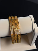 Set of gold bracelets with intricate designs on a white stand against a dark background