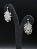 Pair of silver-tone oval filigree drop earrings with scalloped borders and openwork botanical pattern, displayed on a black velvet earring stand