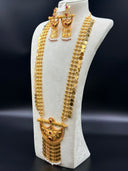 Close-up of a pair of gold-tone chandelier earrings with crescent-shaped bodies, ruby-red and green kundan stones, engraved floral motifs, gold bar tassel drops, and white pearl with red bead finishes, displayed on a cream jewelry bust